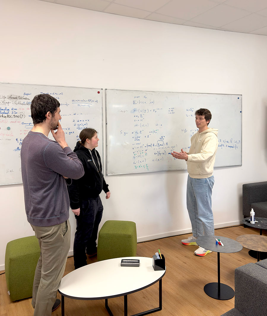 Three people are engaged in conversation in front of two whiteboards covered with equations, while markers and an eraser rest on nearby tables.